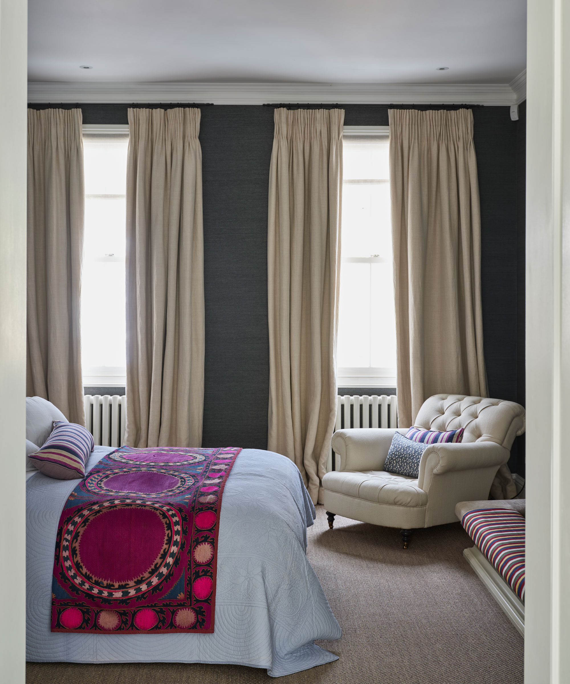 Dormitorio acogedor con paredes pintadas de gris oscuro, cortinas color crema, cama con sábanas blancas y manta rosa, sillón color crema