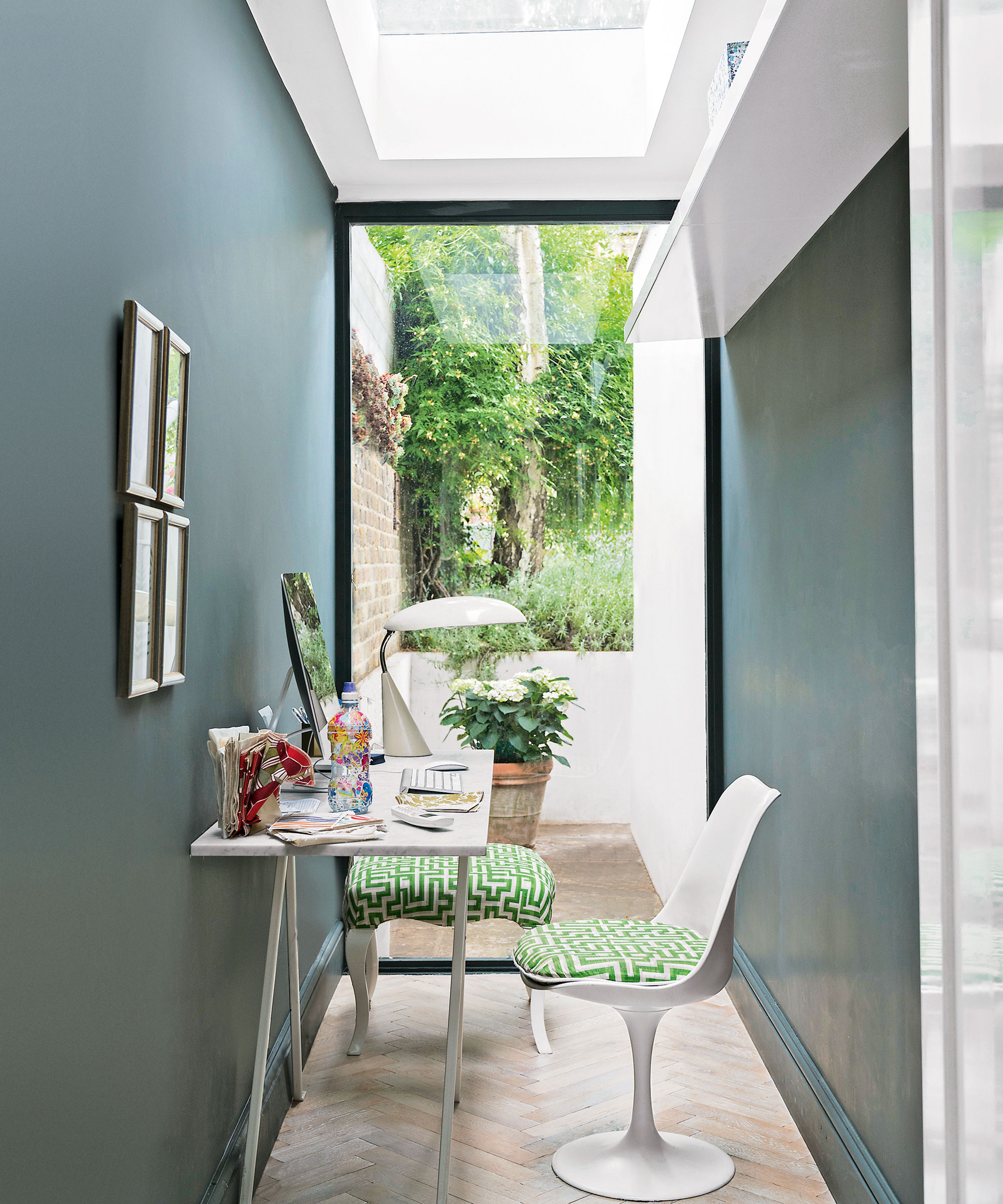 Área de estudio estrecha con piso de parquet, paredes gris azul oscuro, pared de vidrio hacia el jardín, escritorio blanco pequeño y silla tulipán Eero Saarinen con cojín verde.