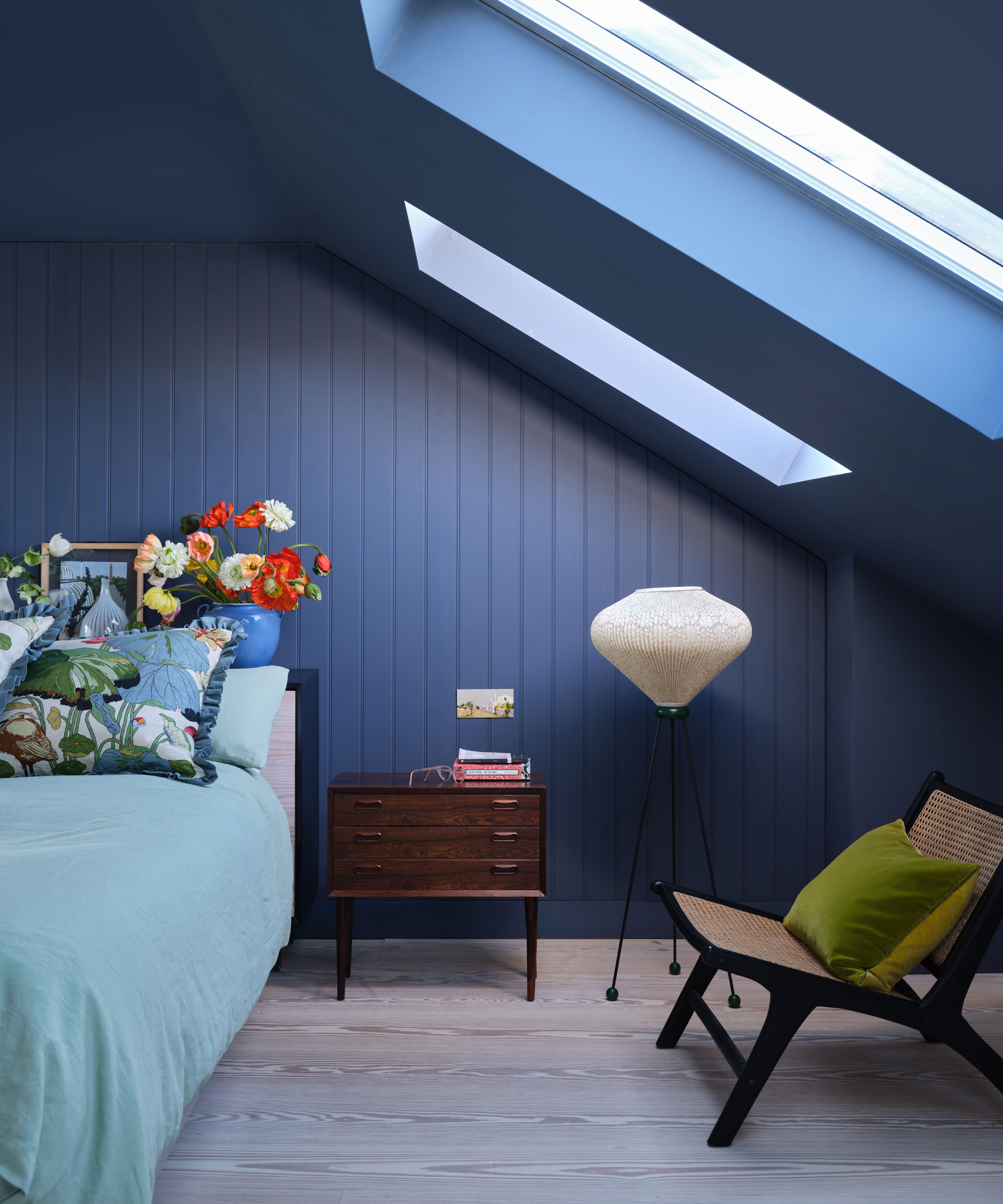 Dormitorio empapado de color pintado de azul profundo, techo inclinado con tragaluces, lámpara de pie, sillón, mesita de noche