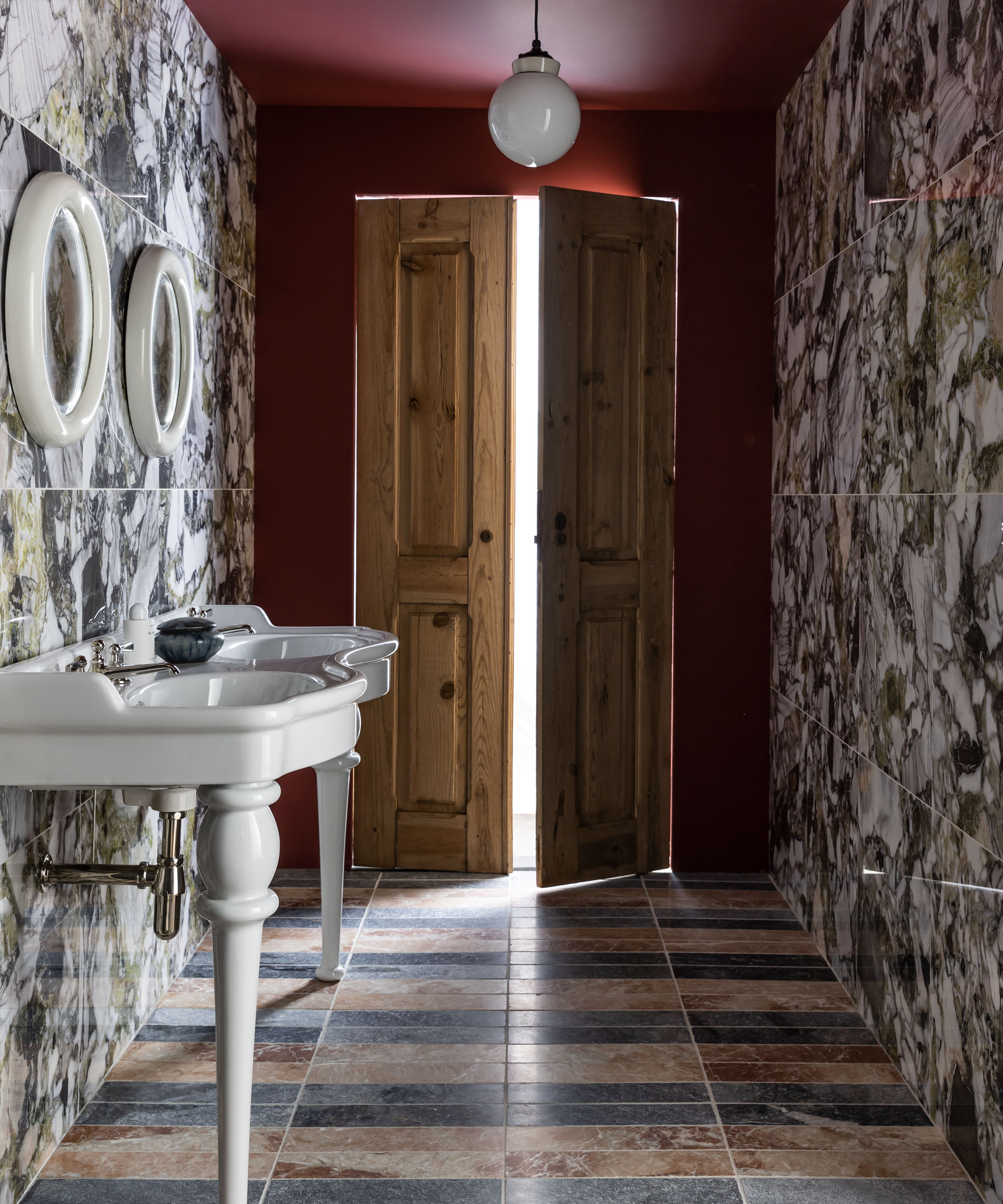 Baño con paredes y techo pintados de rojo, pisos y azulejos de mármol, lavabo doble blanco, espejos blancos redondeados a juego en la parte superior, puertas de madera, colgante redondeado de vidrio blanco