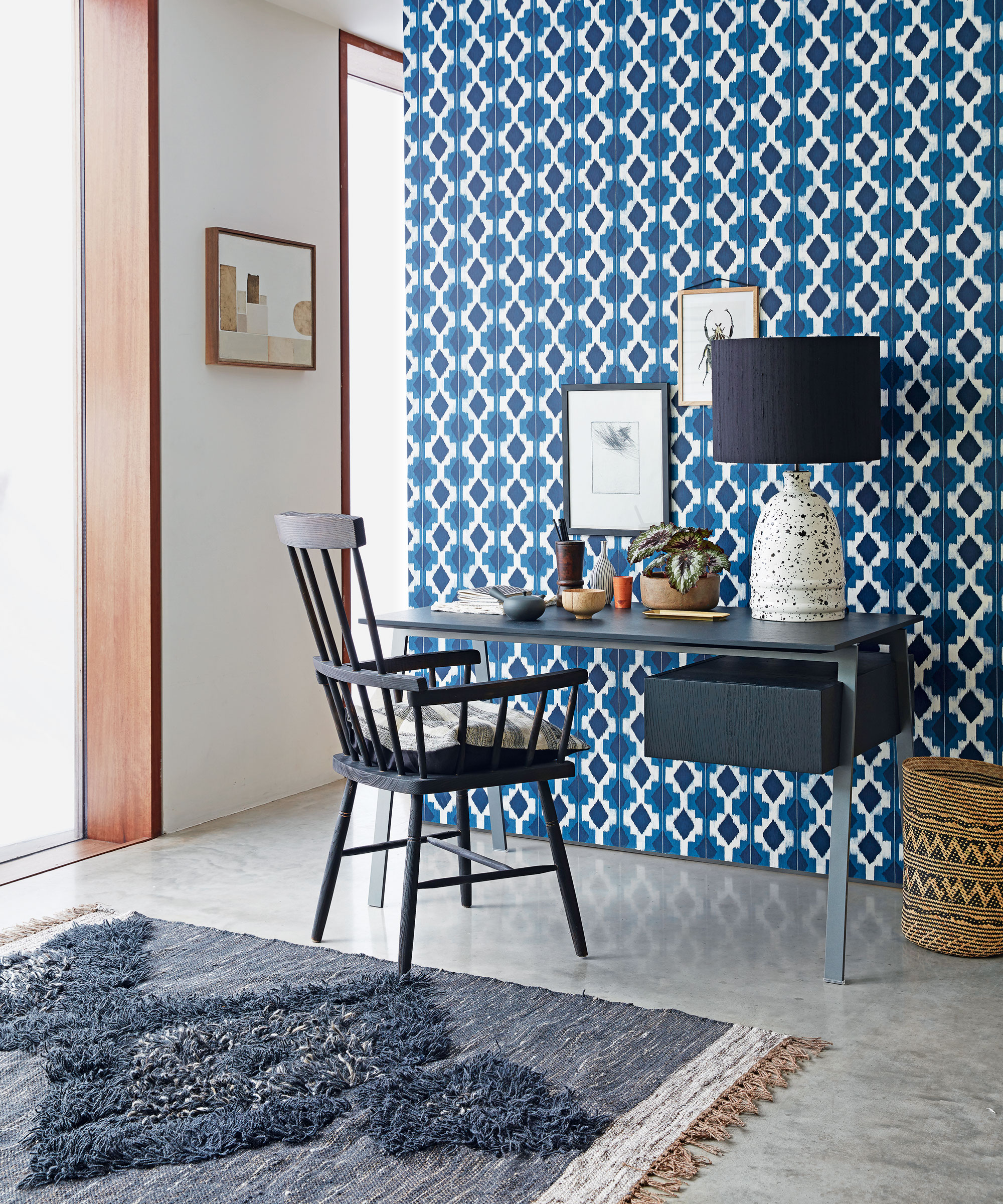 Una oficina en casa con una pared característica empapelada con un patrón geométrico, con suelo de baldosas y alfombra, escritorio y sillón.
