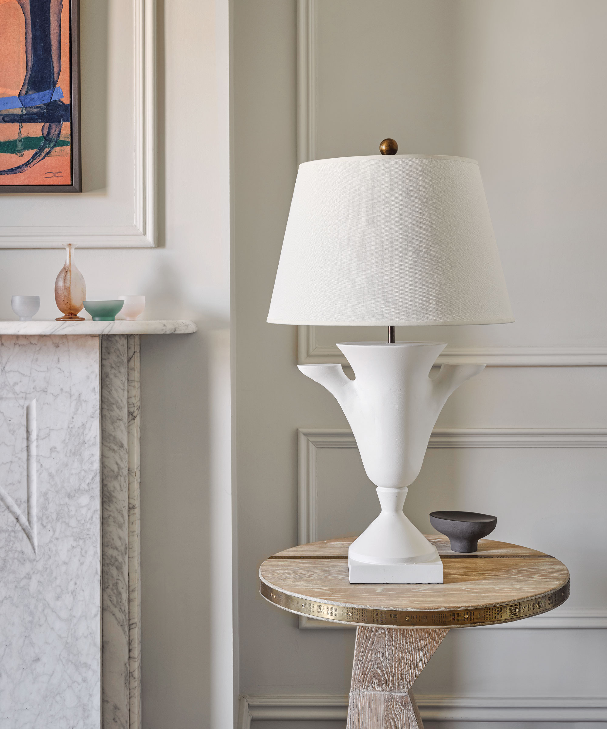 Primer plano de una lámpara de mesa de color crema con una base escultórica de cerámica, colocada encima de una mesa auxiliar de madera, obras de arte y una chimenea en el fondo