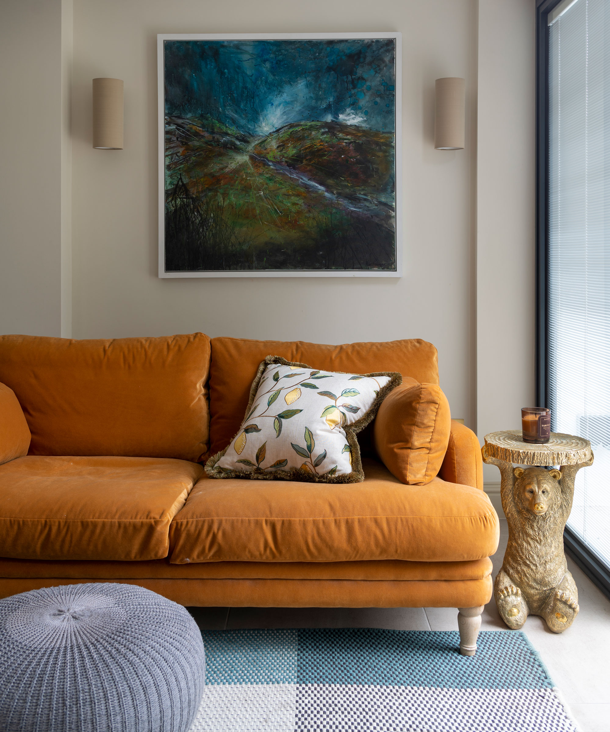 Sala de estar con un acogedor sofá naranja, obras de arte en la pared de atrás, una mesa auxiliar, una otomana redonda y una alfombra azul con motivos cuadrados