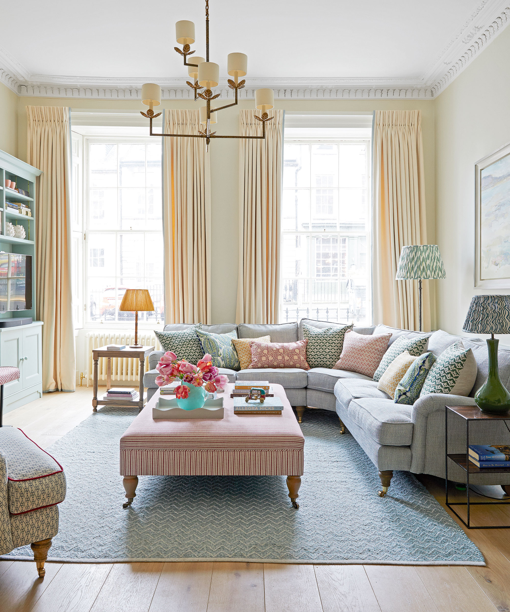 Elegante sala de estar con sofá esquinero gris, cortinas color crema y paredes color crema, alfombra azul, gran otomana estriada, lámpara de araña
