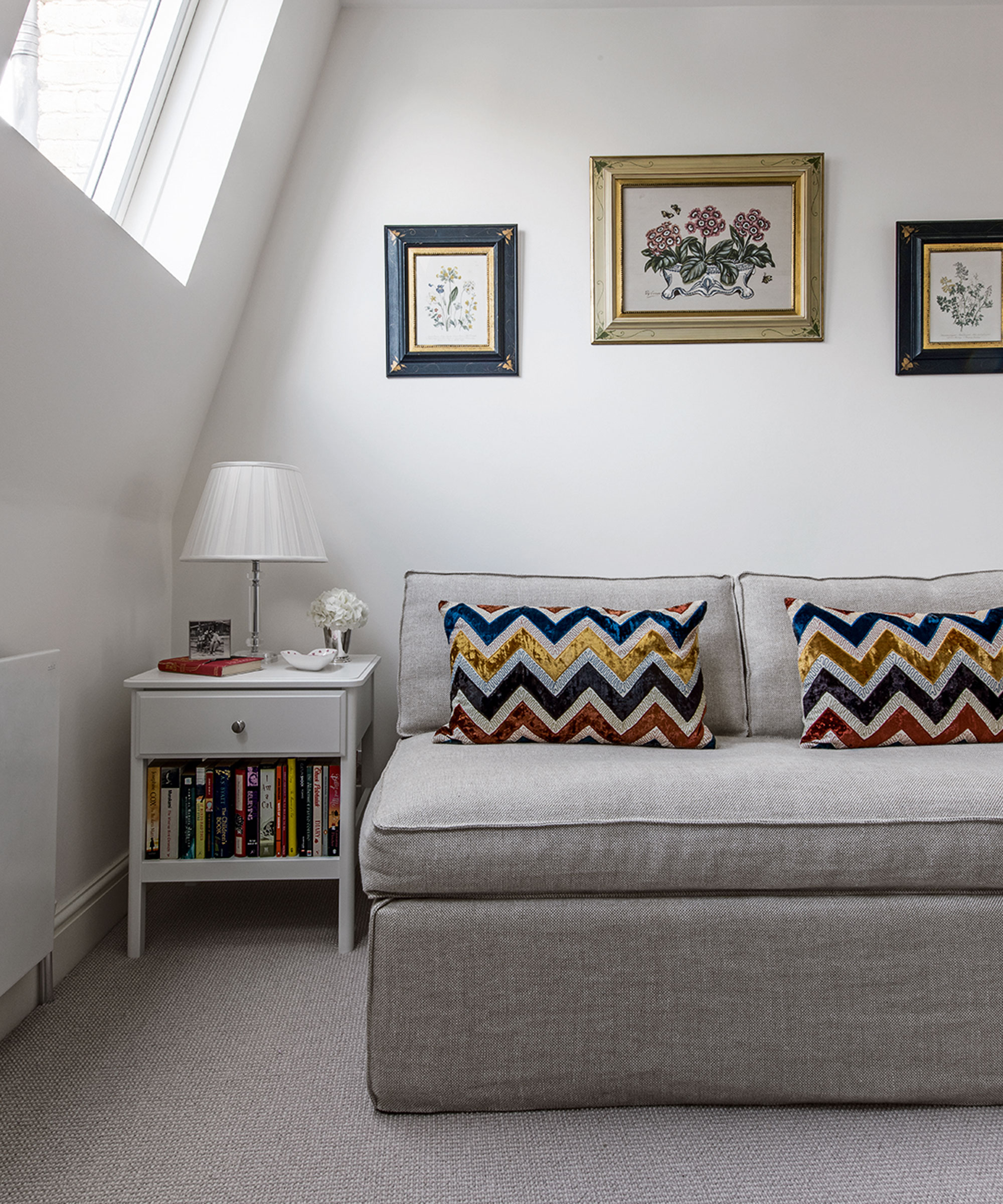 Un dormitorio con un sofá cama gris y cojines estampados en zigzag, aleros inclinados y ventana abuhardillada.