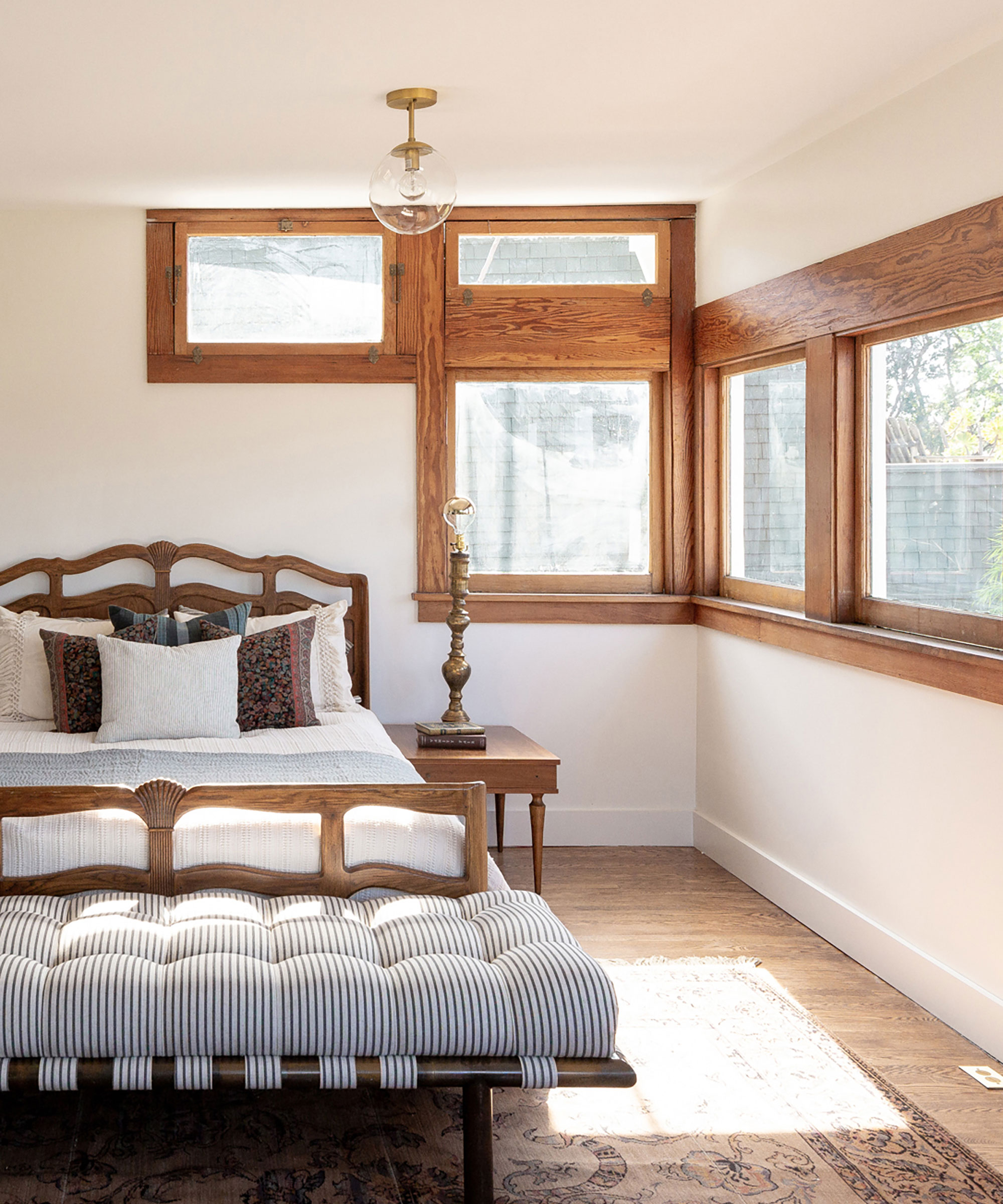 Dormitorio con marcos de ventanas tradicionales de madera, marco de cama de madera oscura con ropa de cama gris, banco y pie de cama